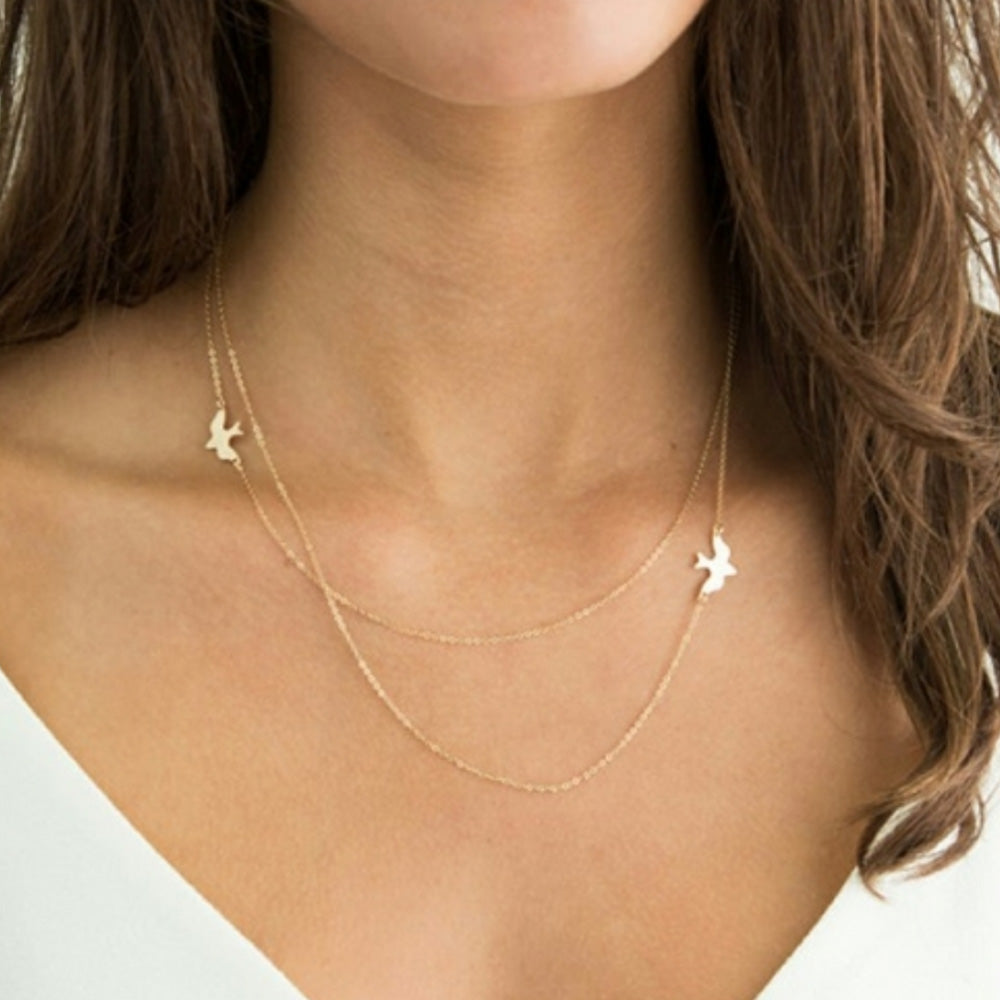 The gold Kelabu double layer necklace with beautiful bird charm worn by a woman with long brown curled hair wearing a white t-shirt